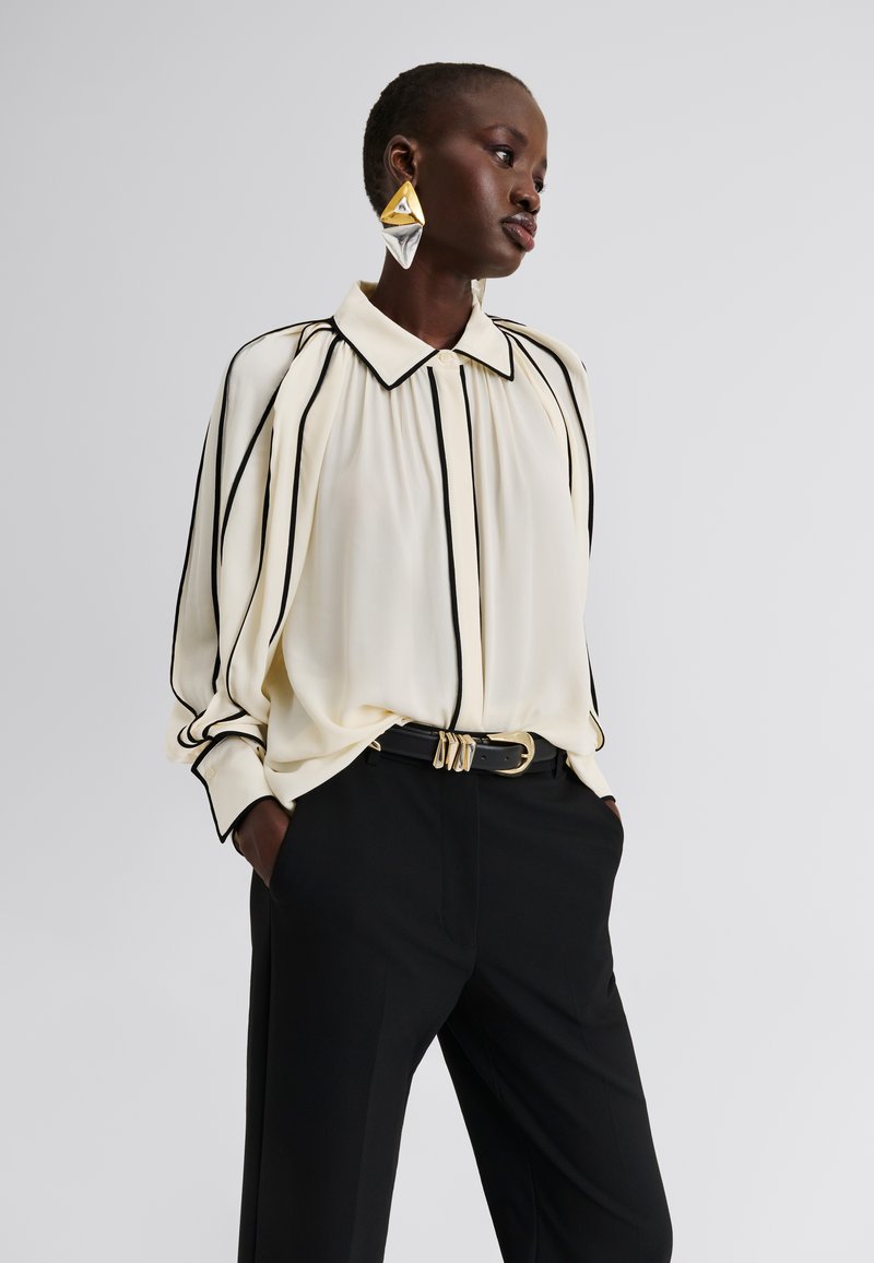 Κρεμ μπλούζα με μαύρες κάθετες ρίγες, γιακά και μανίκια καμπάνα· συνδυασμένη με μαύρο παντελόνι και ζώνη με χρυσές λεπτομέρειες.