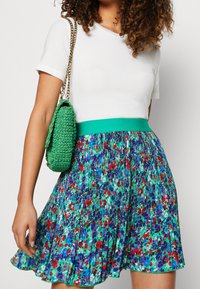 Falda plisada floral con patrones verdes, morados y rojos, que presenta una cinturilla ancha en color azul verdoso. Acompañada de un bolso texturizado verde.
