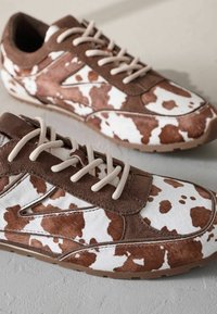 Kuvioituneet lenkkarit, joissa on ruskea ja valkoinen lehmänturkisdesign, mokkaakcentit ja beige nauhat, asetettuna teksturoitua harmaata taustaa vasten.