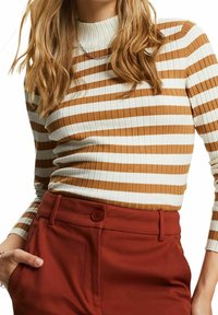 Femme portant un pull à manches longues côtelé avec des rayures horizontales blanches et marron, rentré dans un pantalon taille haute de couleur rouille.