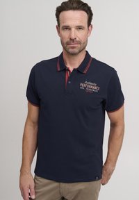 Navy majica s polo ovratnikom, crvenim akcentima, kratkim rukavima i vezenim tekstom "Autentična izvedba". Glatka tekstura, klasičan dizajn ovratnika.