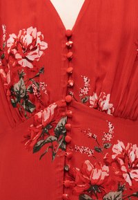 Κόκκινο φόρεμα με κουμπιά καλυμμένα με ύφασμα κατά μήκος του κέντρου, με μεγάλα λουλουδάτα σχέδια σε αποχρώσεις ροζ, λευκού και πράσινα φύλλα.