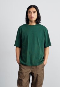 Grøn bomulds-T-shirt med rund hals og korte ærmer, featuring et lille præget logo i centrum. Sammen med brune bukser.