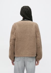 Bruine fleecejas met een gestructureerd oppervlak, verlaagde schouders en geribde zoom. Gecombineerd met lichtgrijze spijkerbroek, gezien van achteren.