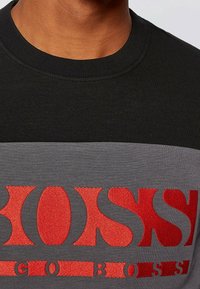 Felpa nera e grigia con scollo rotondo, caratterizzata da un prominente logo rosso ricamato sul petto. Tessuto morbido con una texture liscia.