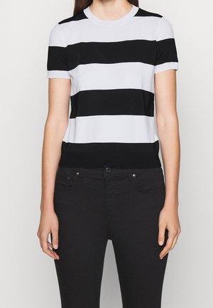 Žena v krátkém rukávu s černo-bílým horizontálně pruhovaným tričkem a černých kalhotách s vysokým pasem, stojící před jednobarevným pozadím.