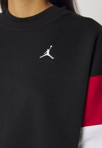 Sudadera negra con cuello de canalé, que presenta el logo Jumpman en blanco y mangas en rojo y blanco con un diseño audaz. Tejido suave con costuras visibles.