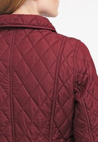 Bordeauxrode doorgestikte jas met een ruitpatroon, opstaande kraag en zichtbare naden, met een gladde textuur en een gewatteerd silhouet.