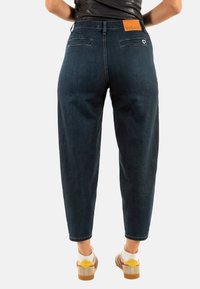 Jeans de mezclilla azul oscuro con un ajuste relajado, bolsillos traseros y una etiqueta de cuero marrón con la inscripción "PLEASE". Combinados con zapatillas multicolores.