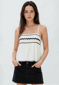 Top bianco con spalline sottili e orlo ondulato nero, abbinato a una gonna di jeans nera con dettagli borchiati e una tasca frontale.