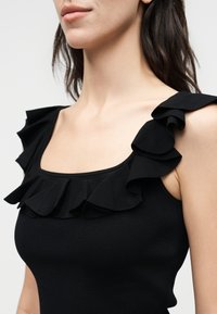 Γυναίκα που φοράει μαύρο αμάνικο τοπ με βολάν στα λουριά και στο ντεκολτέ, εμφανίζοντας τον λαιμό, τους ώμους και το πάνω μέρος του στήθους.