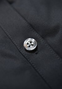 Zwarte stoffen blouse met een opvallende zilveren knop met drie gaten, een gestructureerd oppervlak en zichtbare stiksels.