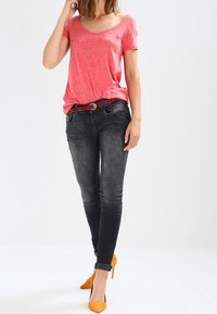 Camiseta de manga corta color coral, jeans ajustados negros y zapatos de tacón amarillo. El conjunto presenta un corte relajado con una textura suave y detalles sutiles.