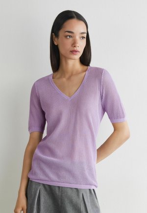 Ung kvinde med glat mørkt hår, iført en lyserød kortærmet V-hals striktop og gråt plisseret skørt, står op ad en hvid væg.