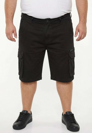 Hombre que lleva pantalones cortos cargo negros con bolsillos laterales y zapatillas negras, de pie frente a un fondo blanco.