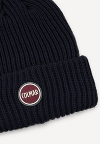 Tumedast kootud beanie, millel on sooniktekstuur, ja ringikujuline burgundia punane silt, millel on valge tekst "COLMAR". Tavaline krae disain.