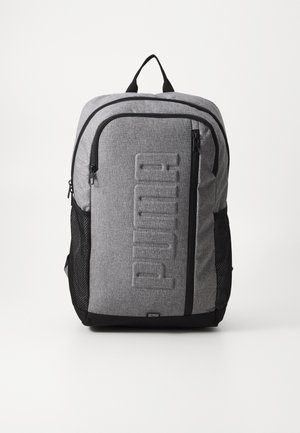 Sac à dos en tissu gris avec une poche zippée à l'avant, des poches latérales en maille et un logo "PUMA" en relief. Il dispose d'une fermeture zippée double et d'accents noirs.