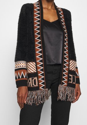Personne portant un cardigan noir duveteux avec un motif géométrique marron, blanc et noir et des franges sur un caraco en satin noir et un pantalon noir.