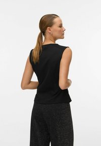 Crna majica bez rukava s strukturiranim dizajnom, koja ima visoki ovratnik, ramene šavne i glatku teksturu. Nosila se uz tamne, teksturirane hlače.
