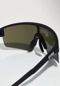 Gafas de sol deportivas con marco negro, grandes lentes tintados en verde, almohadillas ajustables para la nariz y detalles en metal plateado.