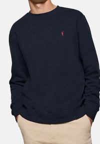 Mörkblå bomullströja med ribbad rund hals, muddar och nederkant. Har en liten röd logotyp på vänster bröst. Slät textur.