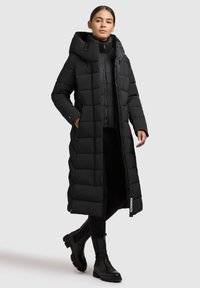 Fekete, hosszú pufertakaró kabát, steppelt dizájnnal, nagy kapucnival és cipzáras záródással. Sötét leggingsek és bokacípők felett viselve. Jellemzője a logó címke.