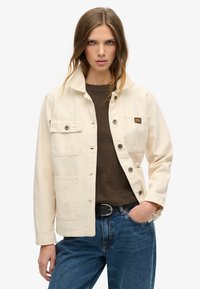 Beige canvasjakke med trykknapper, to brystlommer og en afslappet pasform. Båret over en brun skjorte, parret med blå jeans.