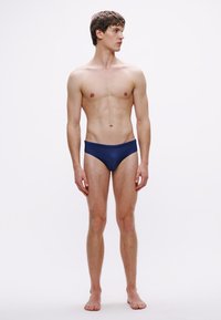 Cuecas de banho azul-marinho feitas de tecido liso, com corte baixo e design minimalista, apresentadas por uma figura masculina num fundo neutro.