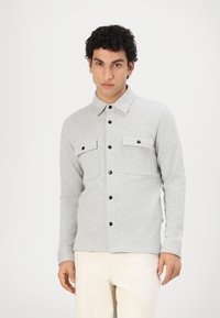 Chemise grise à boutons en tissu doux, avec deux poches poitrine, boutons noirs et col classique. Portée avec un pantalon de couleur claire.