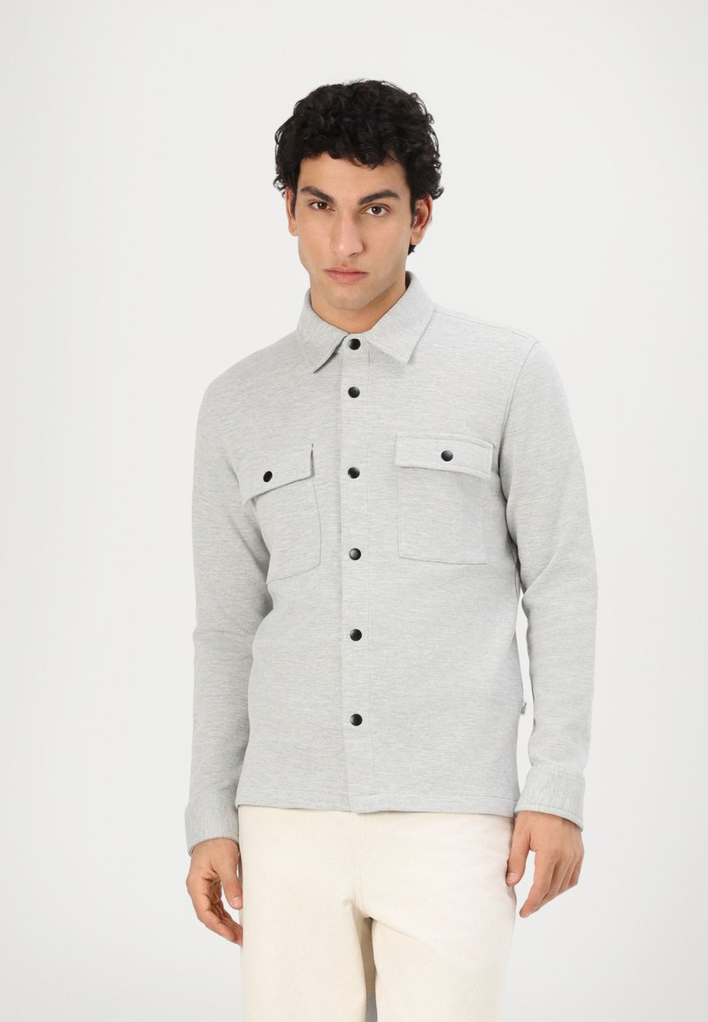 Chemise grise à boutons en tissu doux, avec deux poches poitrine, boutons noirs et col classique. Portée avec un pantalon de couleur claire.