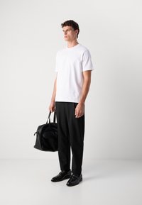 T-shirt blanche à manches courtes, pantalon noir, chaussures en cuir noire, tenant un sac en cuir noir, se tenant devant un fond gris clair.