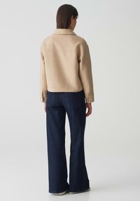 Beige wollen jack met een getailleerde pasvorm, kraag en knoopmanchetten, gecombineerd met donkerblauwe wijde jeans. Soepele textuur.