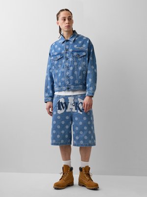 Jeune homme aux cheveux tressés portant une veste en jean bleue à pois et un short assorti à hauteur des genoux avec des bottes de travail couleur tan.