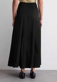 Jupe A-line vert foncé avec une texture lisse, dotée d'une taille ajustée et de drapés subtils. Portée avec des chaussures noires.