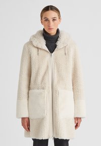 Frau in einem cremefarbenen, strukturierten Fleece-Mantel mit Kapuze, großen Vordertaschen und einem Reißverschluss vorne, über einem dunklen Rollkragenpullover.