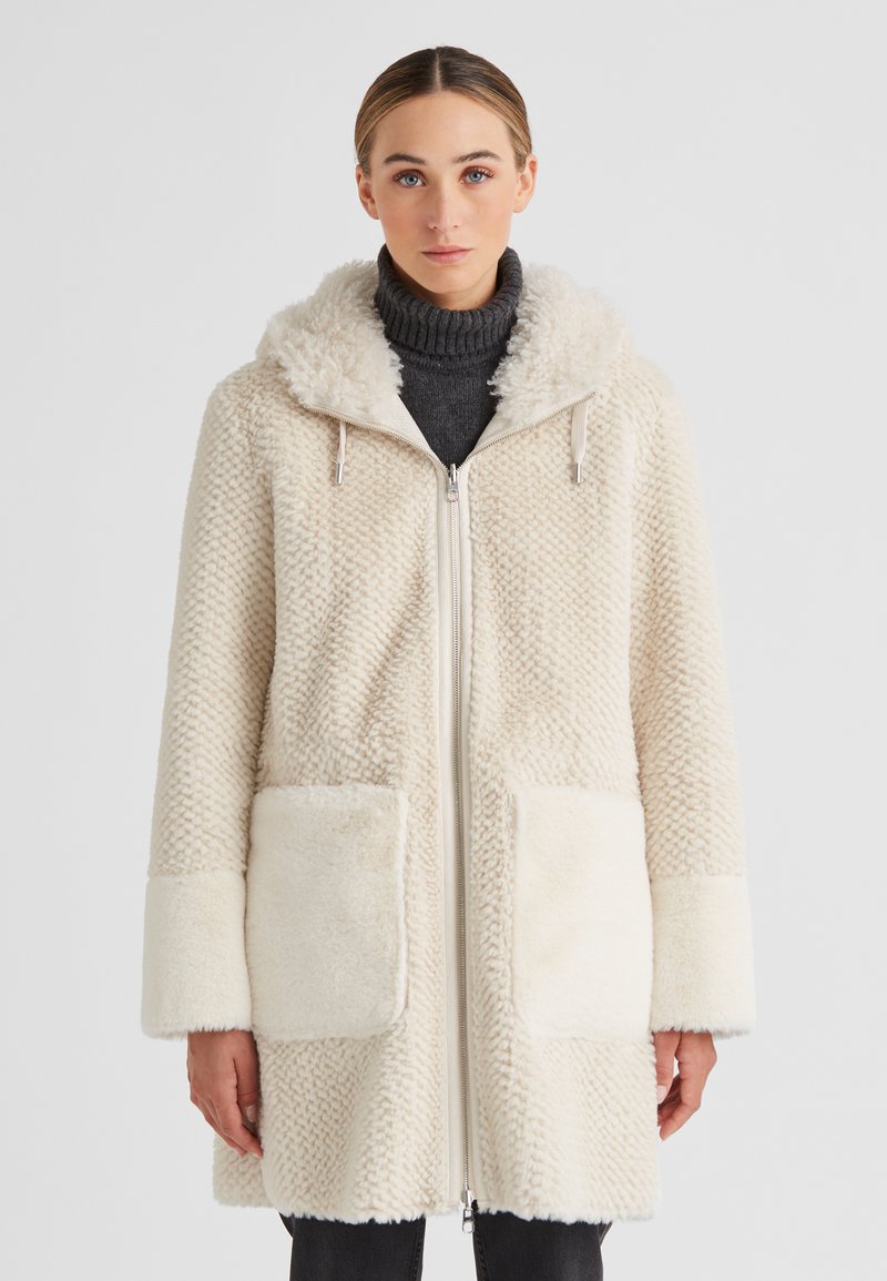 Frau in einem cremefarbenen, strukturierten Fleece-Mantel mit Kapuze, großen Vordertaschen und einem Reißverschluss vorne, über einem dunklen Rollkragenpullover.