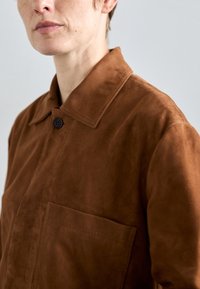 Κοντινό πλάνο ενός ατόμου που φοράει ένα καφέ σουέτ τζάκετ με μεγάλο γιακά, ορατό κουμπί και τσέπη στο στήθος σε ουδέτερο φόντο.