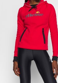 Femme portant un sweat à capuche rouge de la marque ellesse avec des poches zippées noires et des leggings de sport noirs avec des détails imprimés sur les cuisses.