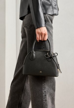 Persoon houdt een kleine zwarte leren handtas met strikdetail vast, draagt een zwarte leren jas en grijze jeans tegen een effen achtergrond.