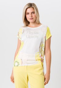 T-shirt léger blanc avec imprimés colorés de fruits, manches courtes, et texte en strass "FRESH LEMON" au centre. Associé à un pantalon jaune.