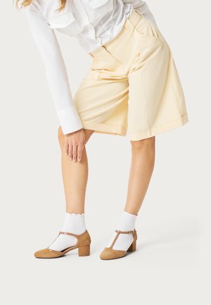 Vrouw met beige ruime korte broek, wit shirt met lange mouwen, witte sokken en bruine T-strap schoenen met blokhak, die naar voren buigt en haar been aanraakt.