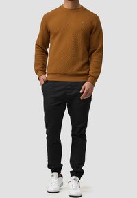 Brązowa bluza z wzorem, z okrągłym dekoltem, teksturowanym materiałem, zestawiona z czarnymi spodniami i białymi trampkami. Prosty design, z elastycznymi mankietami.