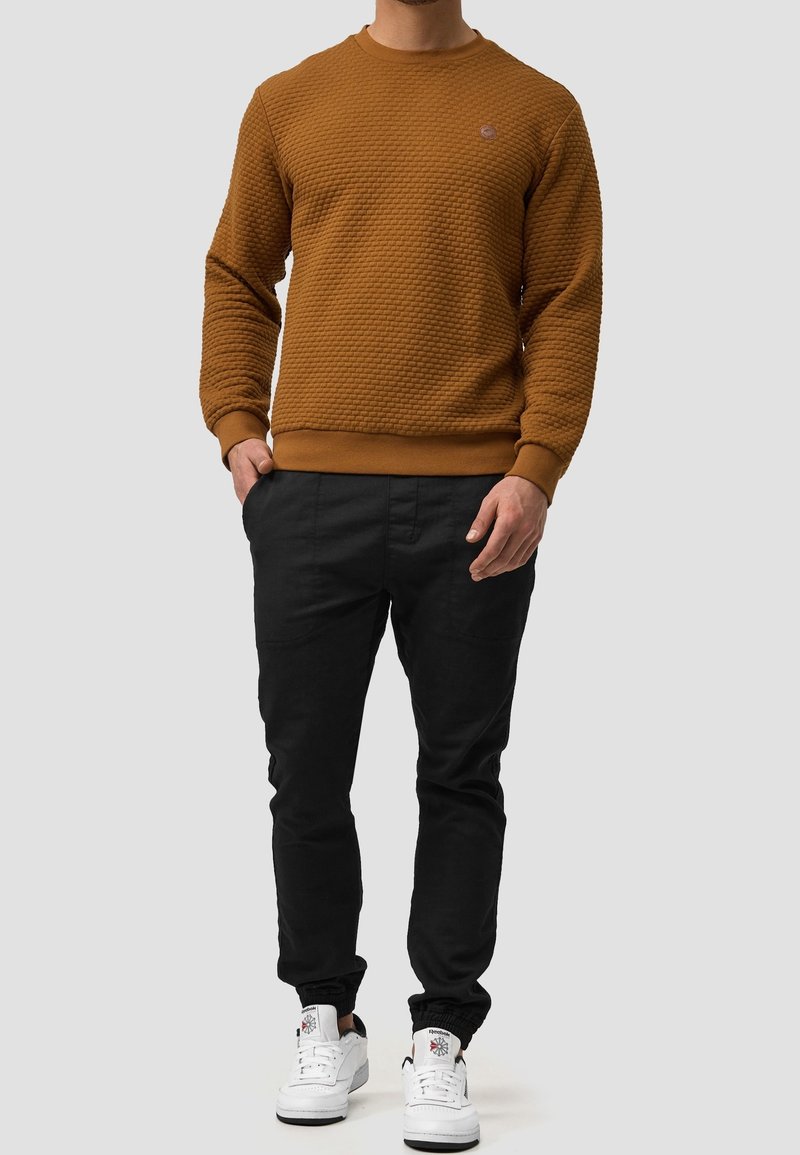 Brązowa bluza z wzorem, z okrągłym dekoltem, teksturowanym materiałem, zestawiona z czarnymi spodniami i białymi trampkami. Prosty design, z elastycznymi mankietami.