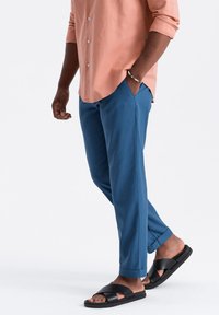 Pantalon bleu avec une coupe ajustée, associé à une chemise à boutons pêche. Sandales noires avec un design à lanières croisées. Tenue simple et décontractée.