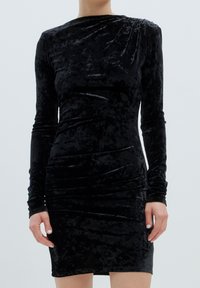 Schwarzes Samtkleid mit langen Ärmeln, das eine taillierte Silhouette, geraffte Seitendetails und einen runden Ausschnitt aufweist, wodurch eine glatte, weiche Textur entsteht.
