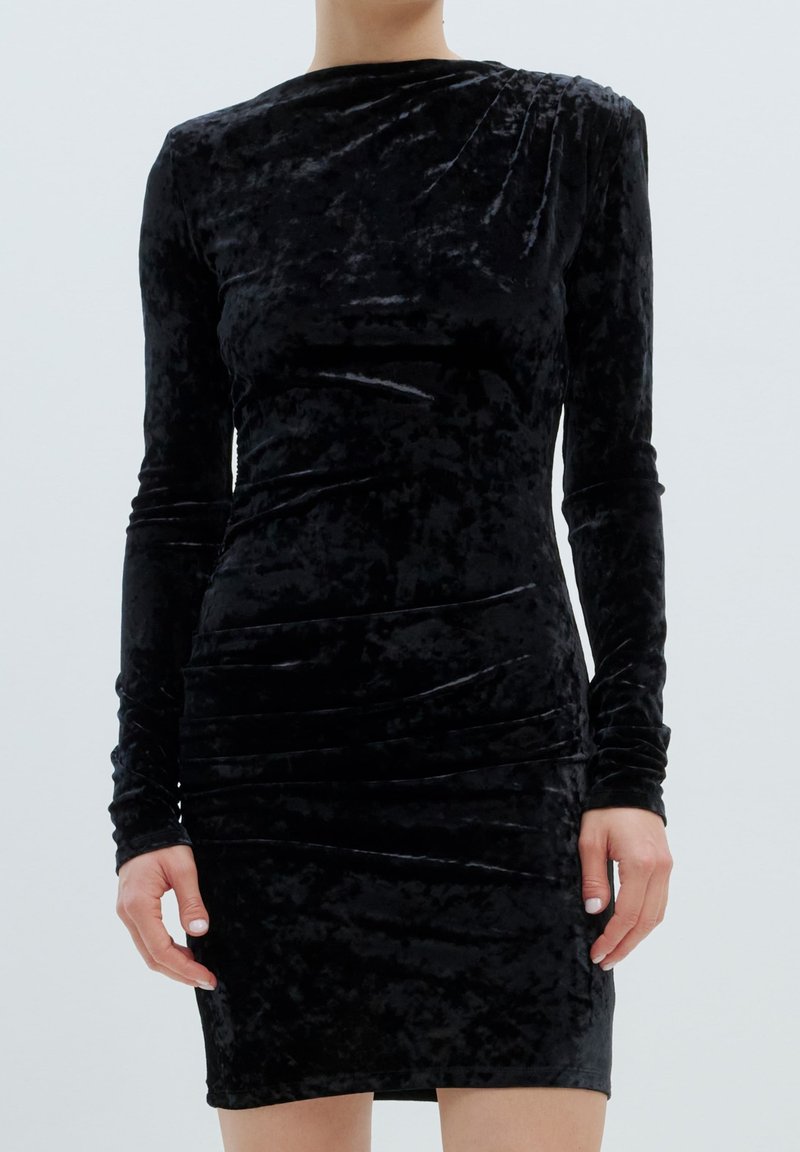 Schwarzes Samtkleid mit langen Ärmeln, das eine taillierte Silhouette, geraffte Seitendetails und einen runden Ausschnitt aufweist, wodurch eine glatte, weiche Textur entsteht.