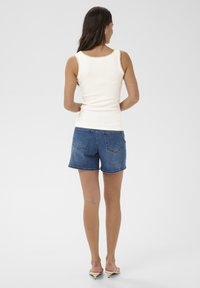 Mujer de espaldas, vistiendo una blusa blanca sin mangas, shorts de mezclilla azules y sandalias beige de tacón bajo sobre un fondo liso.