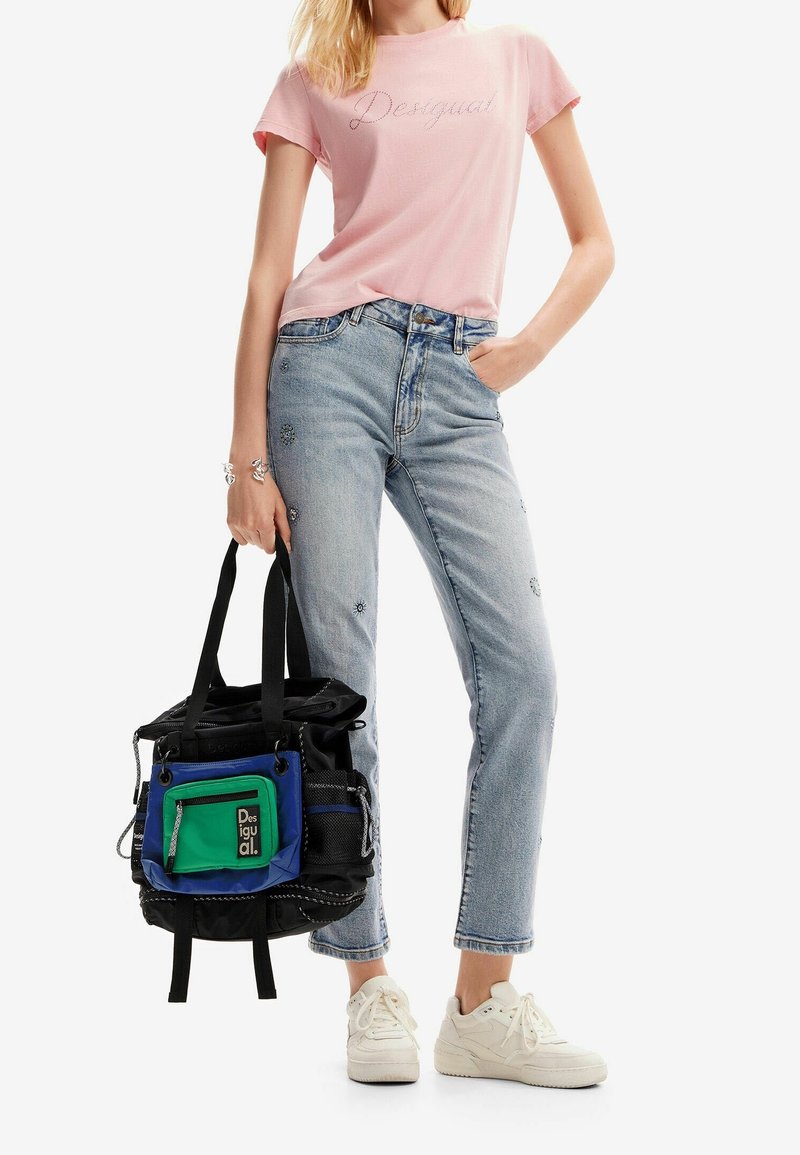 Femme en t-shirt rose clair et jean bleu orné, tenant un sac cabas Desigual noir, bleu et vert, portant des baskets blanches.