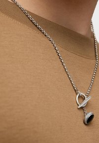 Collana in argento con ciondolo circolare dotato di una pietra nera e un anello decorativo, indossata sopra una maglietta color beige con colletto a girocollo.