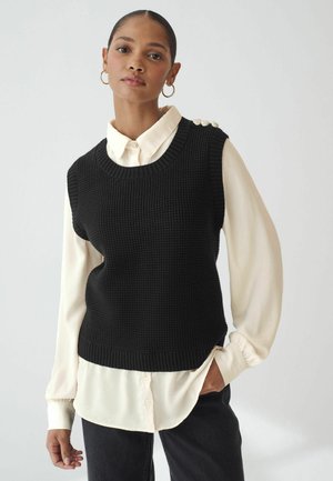 Sort strikket ærmeløs sweater over en hvid skjorte med krave, der har tekstureret stof, ribbet halsudskæring og dekorative knapper på skulderen.